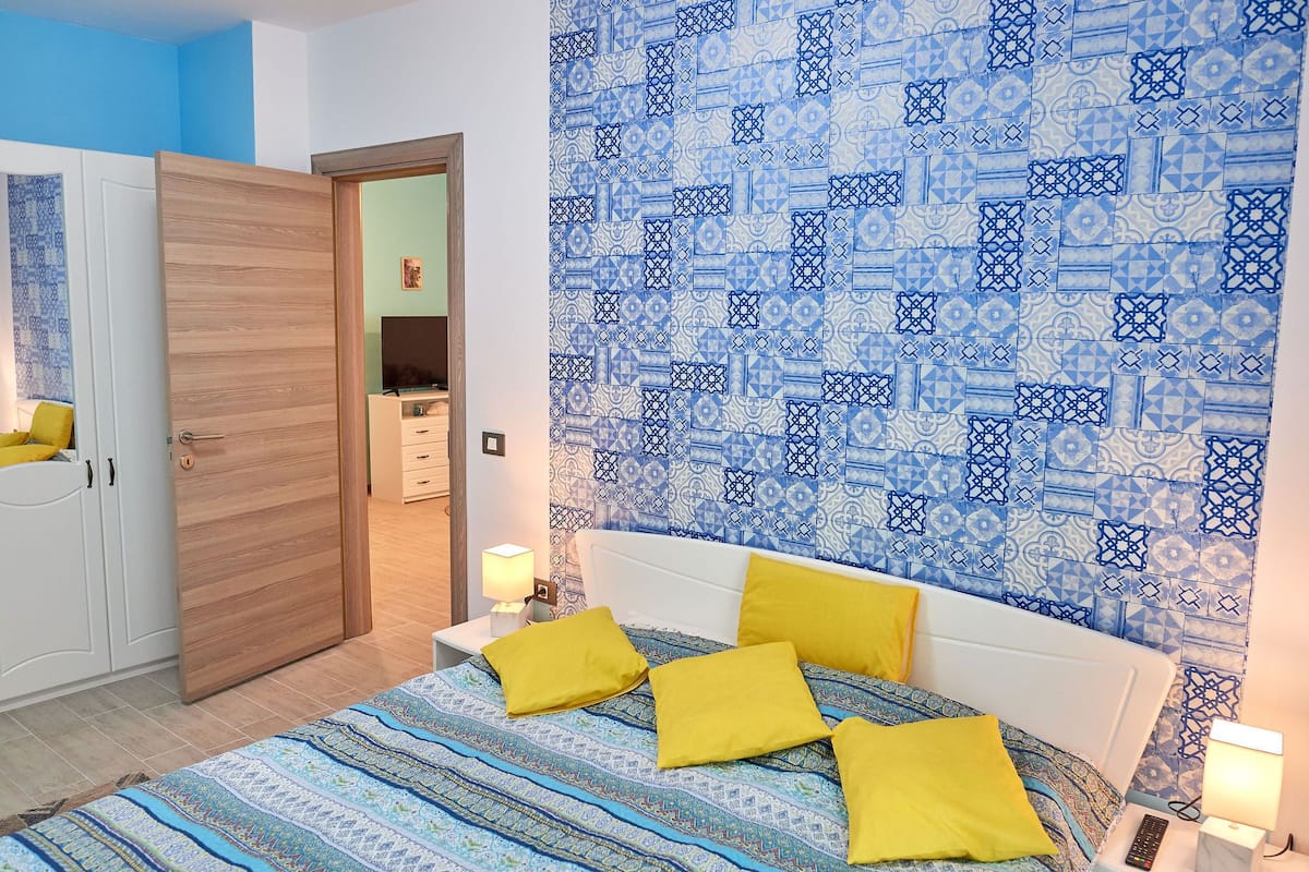 A bedroom with a patterned blue and white wall, a bed with a blue and white patterned bedspread, and yellow pillows.
