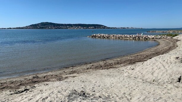 Una playa arenosa con aguas tranquilas, un rompeolas rocoso y una colina distante.