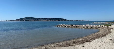 Une plage de sable fin aux eaux calmes, une digue rocheuse et une colline qui se dessine à l’horizon.