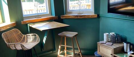 Una habitación con un escritorio de madera, dos sillas, un taburete pequeño, un televisor y una ventana que da a vistas del exterior.