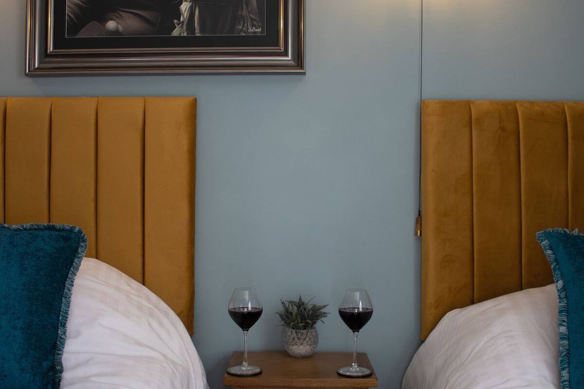 A hotel room with two beds, a nightstand with wine glasses, a framed picture, and a wall-mounted light.