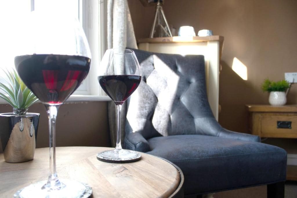 A room with a round wooden table, two wine glasses, a potted plant, and a grey upholstered chair.