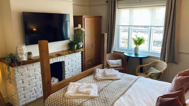 A hotel room with a fireplace, a television, a bed with a quilt, a chair, and a window with curtains. - The Riverside Rooms (Newark)