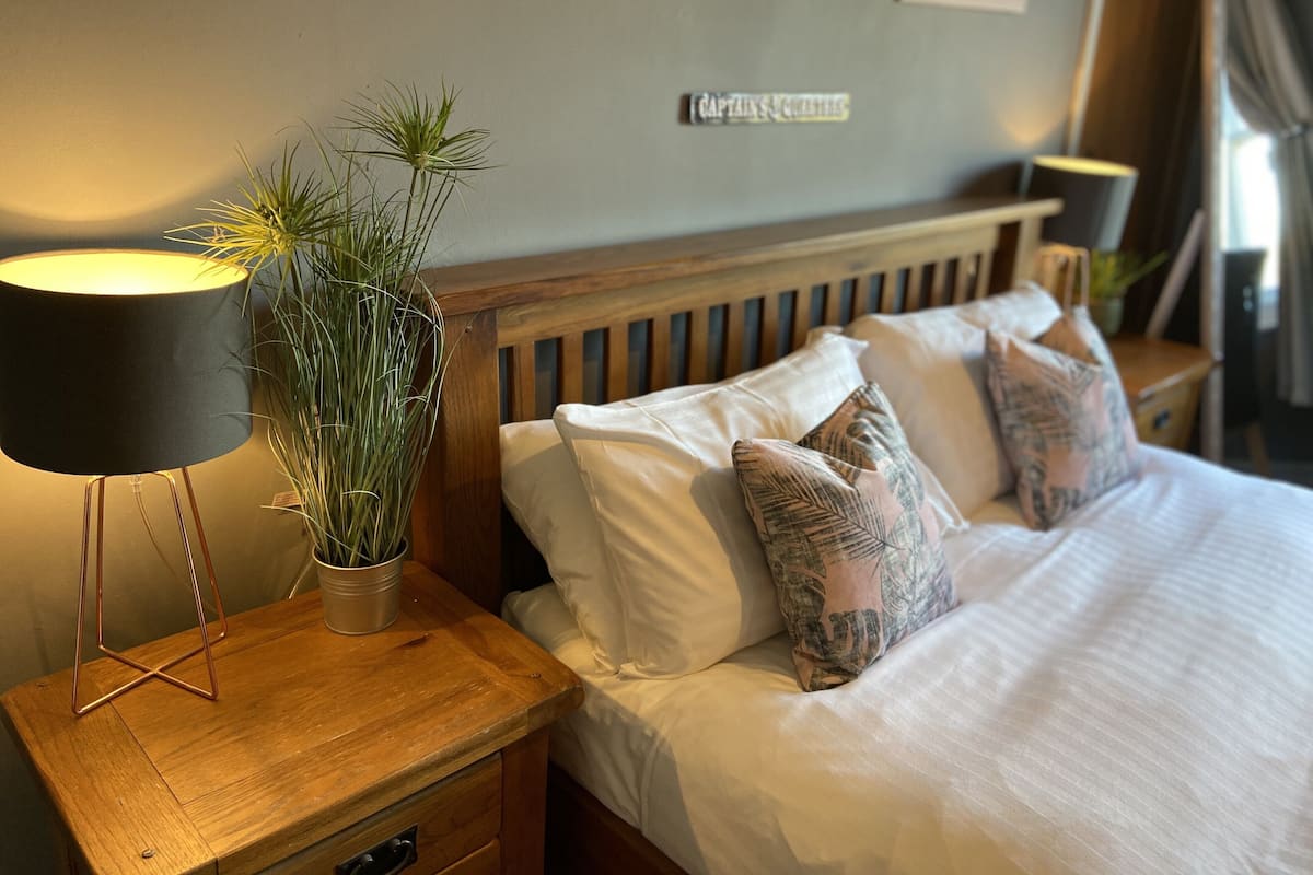 A neatly made bed with white linens and decorative pillows, a bedside lamp, and a potted plant on a wooden nightstand.