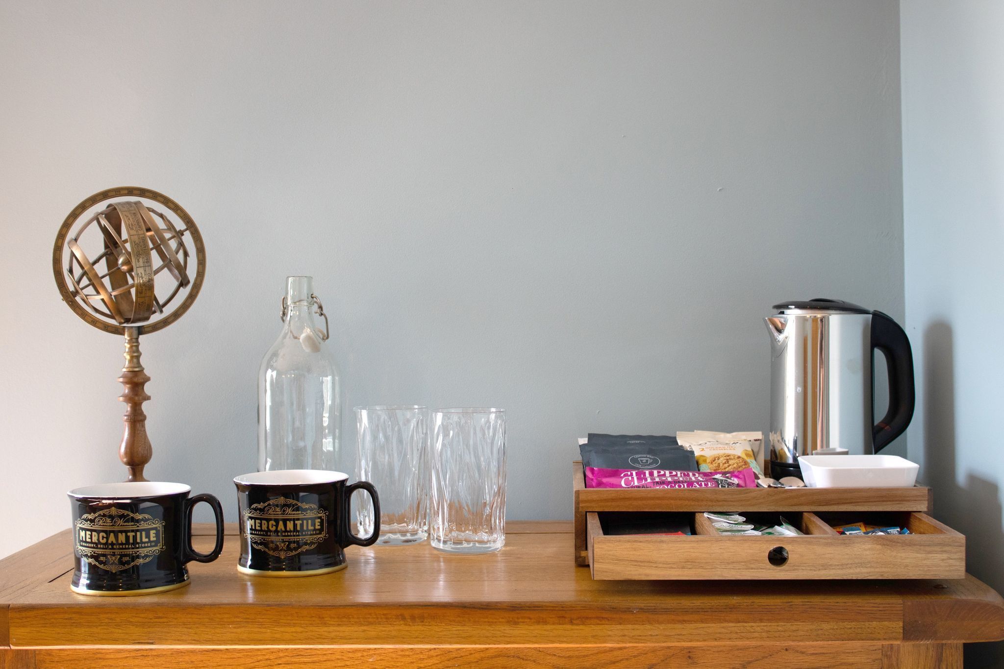 Een houten tafel met twee mokken, een glazen fles, twee glaasjes, een koffiezetapparaat en een doos snacks.