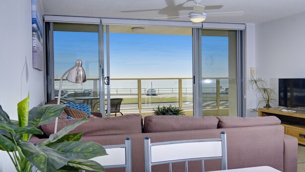 A living room with a sofa, a TV, a ceiling fan, and a view of the beach through large glass doors. - Ebbtide 2 - On Main Beach! (Forster)