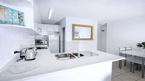 A modern kitchen with white cabinetry, a built-in microwave, a toaster, and a sink. There is a dining area with a table and chairs. - Ebbtide 2 - On Main Beach! (Forster)