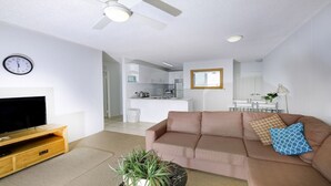 A modern living room with a flat-screen TV, a wooden entertainment unit, a beige sofa with patterned cushions, a potted plant, and a ceiling fan. - Ebbtide 2 - On Main Beach! (Forster)