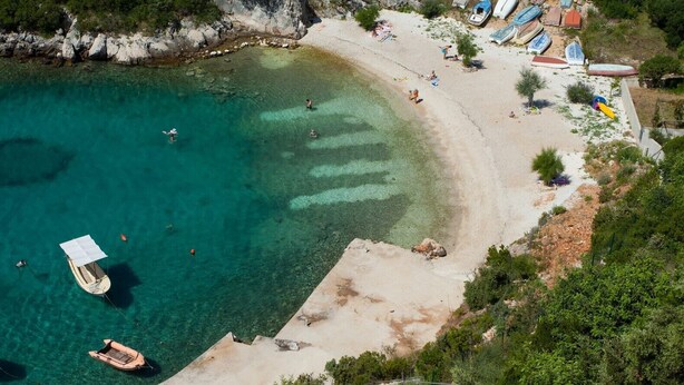 Vue aérienne d’une plage aux eaux turquoise et limpides, avec des bateaux et une rive sablonneuse.