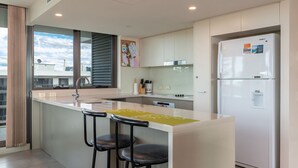 A modern kitchen with a central island, two bar stools, a refrigerator, and a window with blinds.