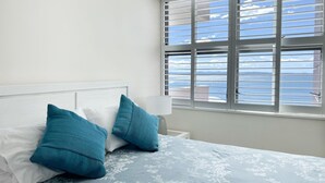 Un dormitorio con una cama, dos almohadas, una mesita de noche, una lámpara y una ventana con persianas.
