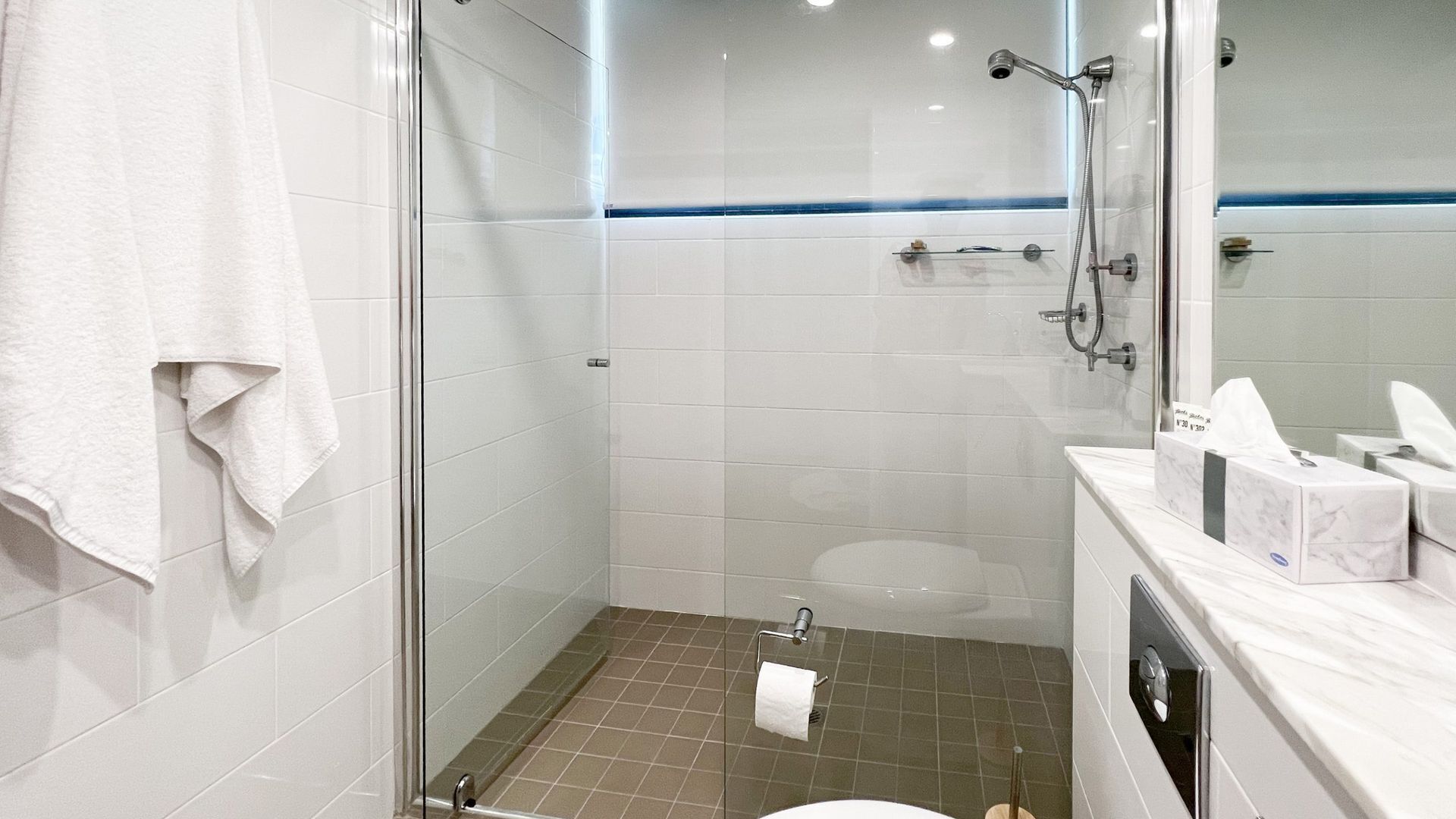 Une salle de bain moderne avec une douche à parois de verre, des carreaux blancs, des toilettes et un meuble-lavabo avec un miroir et des articles de toilette.