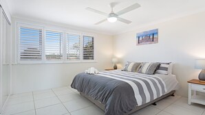 A bedroom with a bed, a ceiling fan, a window with blinds, a bedside table with a lamp, and a painting on the wall.