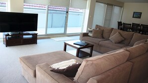 A living room with a large flat-screen TV, a sofa, a coffee table, and a dining area with chairs. - The Reef 103 - Lakeside! (Forster)