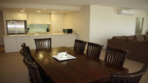 A modern kitchen with white cabinets, a stainless steel refrigerator, and a dining area with a wooden table and chairs. - The Reef 103 - Lakeside! (Forster)