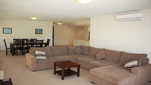 A living room with a sectional sofa, a coffee table, a dining area with a table and chairs, and an air conditioning unit on the wall. - The Reef 103 - Lakeside! (Forster)