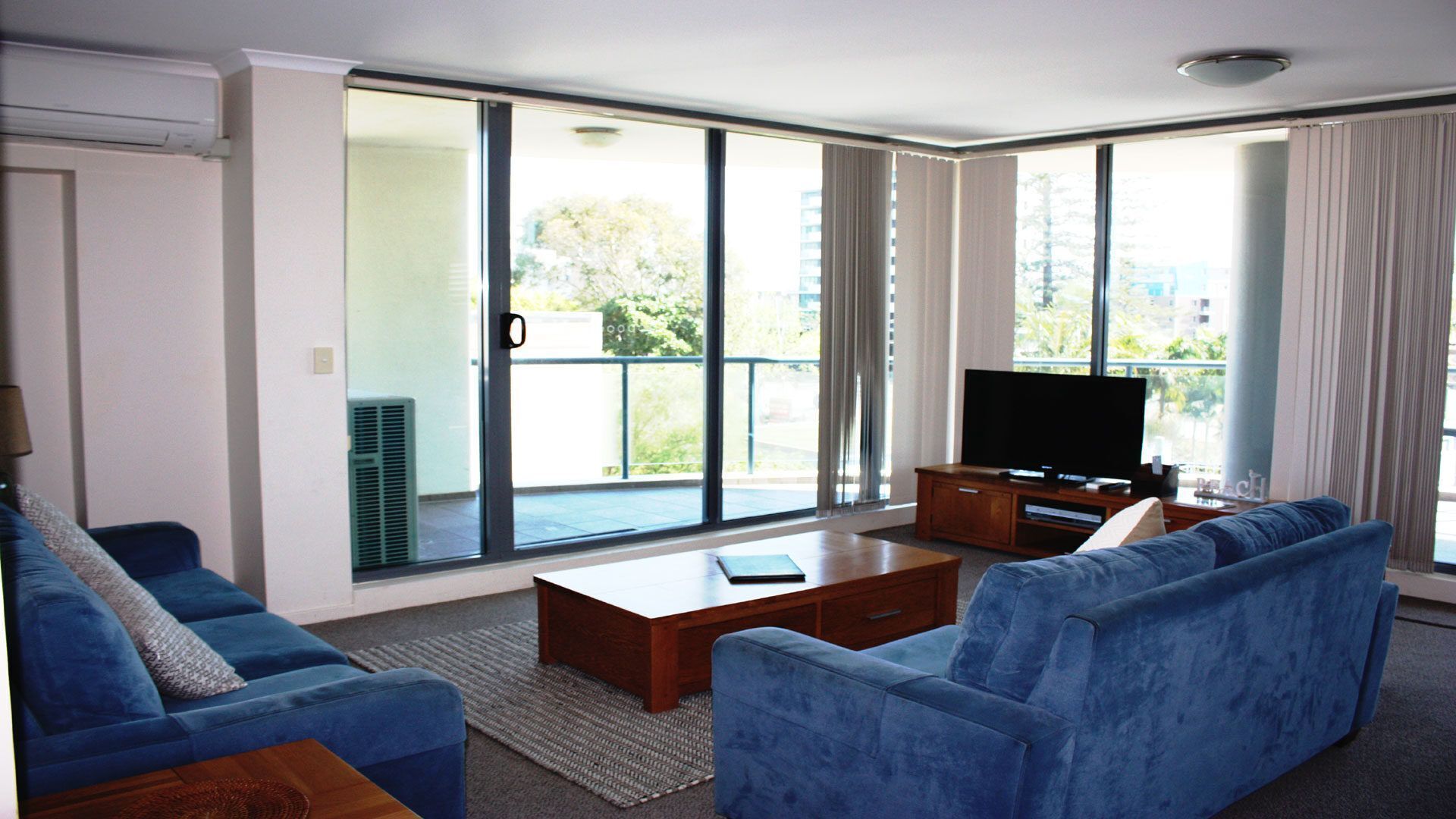 A modern living room with a large sliding glass door leading to a balcony, a flat-screen TV, a blue sofa, and a wooden coffee table.