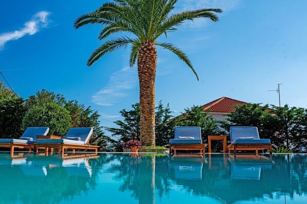 A pool with lounge chairs, a palm tree, and a building with a red roof. - Amor Beach Villa 3 Bedrooms with huge pool, BBQ, near the beach. (Zakynthos)