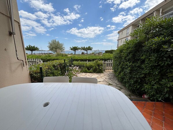 A patio area with a table and chairs, a view of the sea, and a hedge.