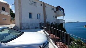 Ein Küstengebäude mit Balkon und Meerblick, ein Auto vor der Fassade sowie eine Treppe zum Eingang.