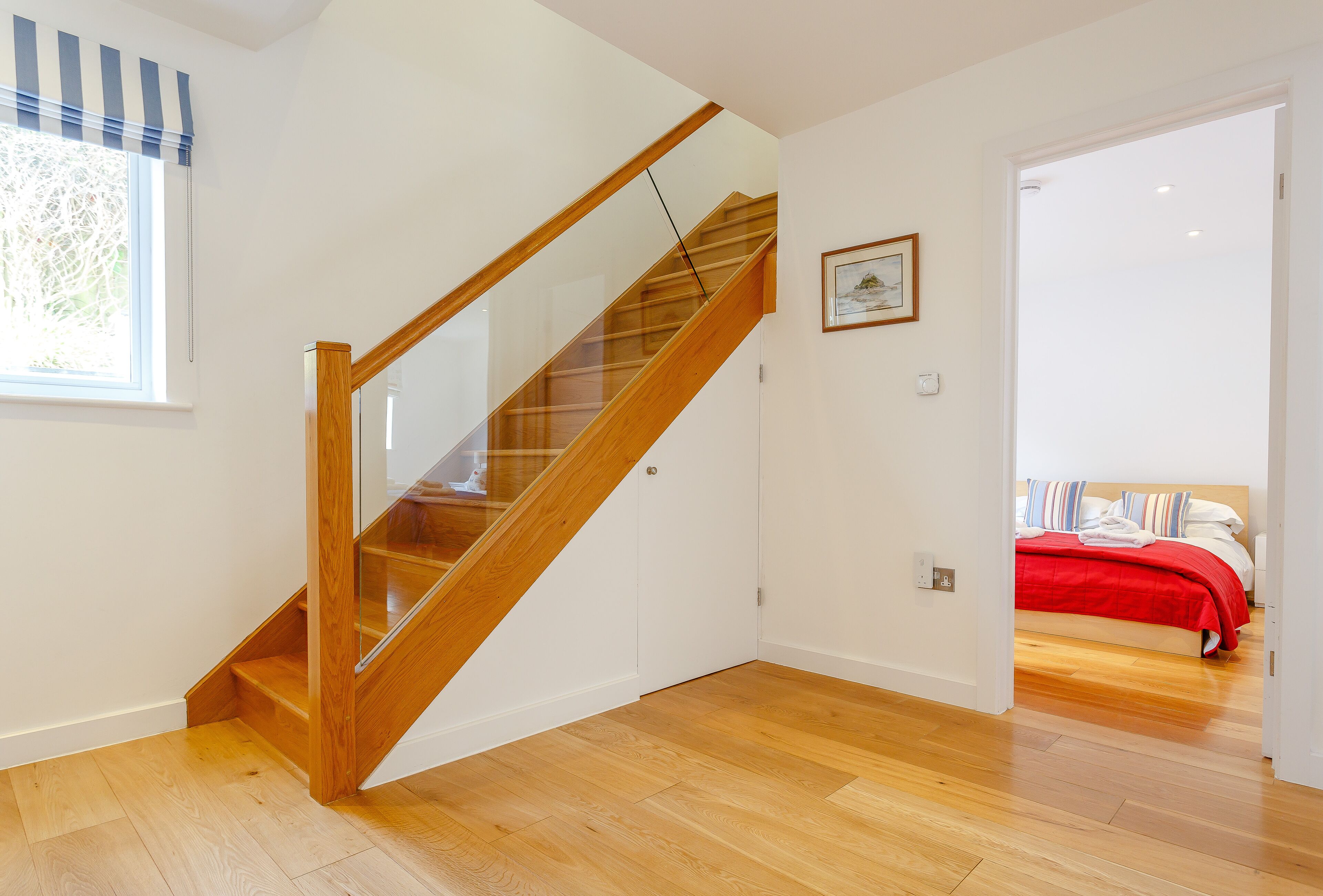 A modern interior with a wooden staircase, a bedroom with a red bedspread, and a framed picture on the wall.