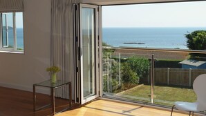 A modern living room with a large sliding glass door leading to a balcony with a sea view. - Luxury property Sleeps 8 - Sea views & hot tub - 200m from beach! (Bigbury-on-Sea)