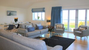A modern living room with a grey sofa, two armchairs, a glass coffee table, and a dining area with a table set for four. - Luxury property Sleeps 8 - Sea views & hot tub - 200m from beach! (Bigbury-on-Sea)