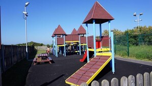 A playground with multiple play structures, including slides and climbing areas, surrounded by a wooden fence and a clear sky. - 1h from central London and 30min from Dover, opposite stairs to sandy beach (Broadstairs)