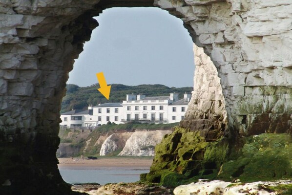 A coastal view through a stone archway, overlooking a white building with multiple windows and a balcony, situated on a cliff with a beach and rocky terrain in the foreground.