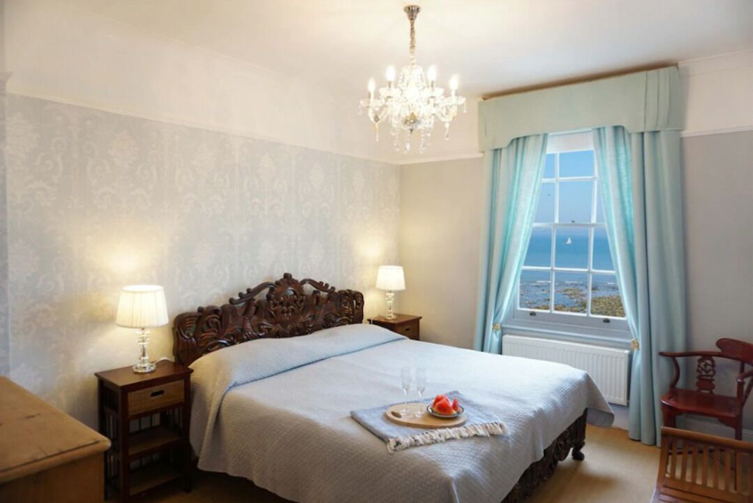 Ein Schlafzimmer mit einem großen Bett, zwei Nachttischlampen, einer Kristallleuchtre und einem Fenster mit Vorhängen, mit Blick auf das Meer.