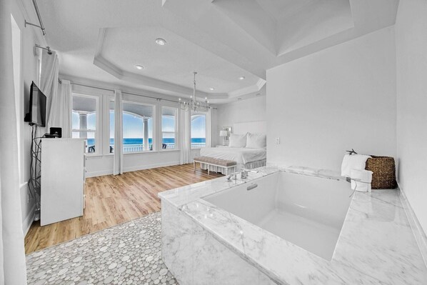Un dormitorio moderno con un ventanal grande, una bañera de mármol y piso de madera.