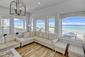 Un salón amplio con un gran sofá beige en forma de L, suelo de madera y ventanas grandes que ofrecen vistas a la playa y al océano.