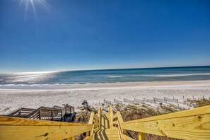 Una playa con un paseo marítimo de madera que conduce a una zona arenosa, cielo azul despejado y el océano al fondo.