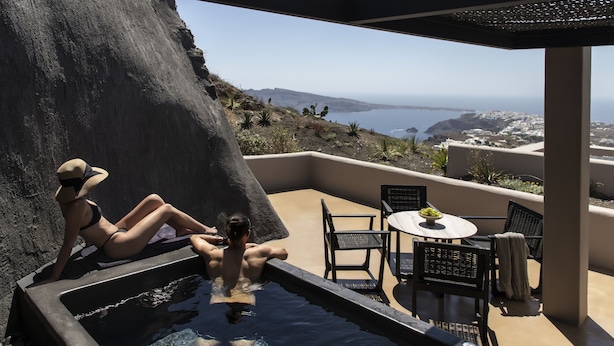 A hot tub with two people, a table with chairs, and a view of the sea and landscape. - Nimbus Santorini (Santorini)