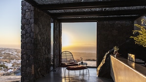 A modern room with a stone wall, a view of the landscape, a wicker chair, and a small table with a vase. - Nimbus Santorini (Santorini)