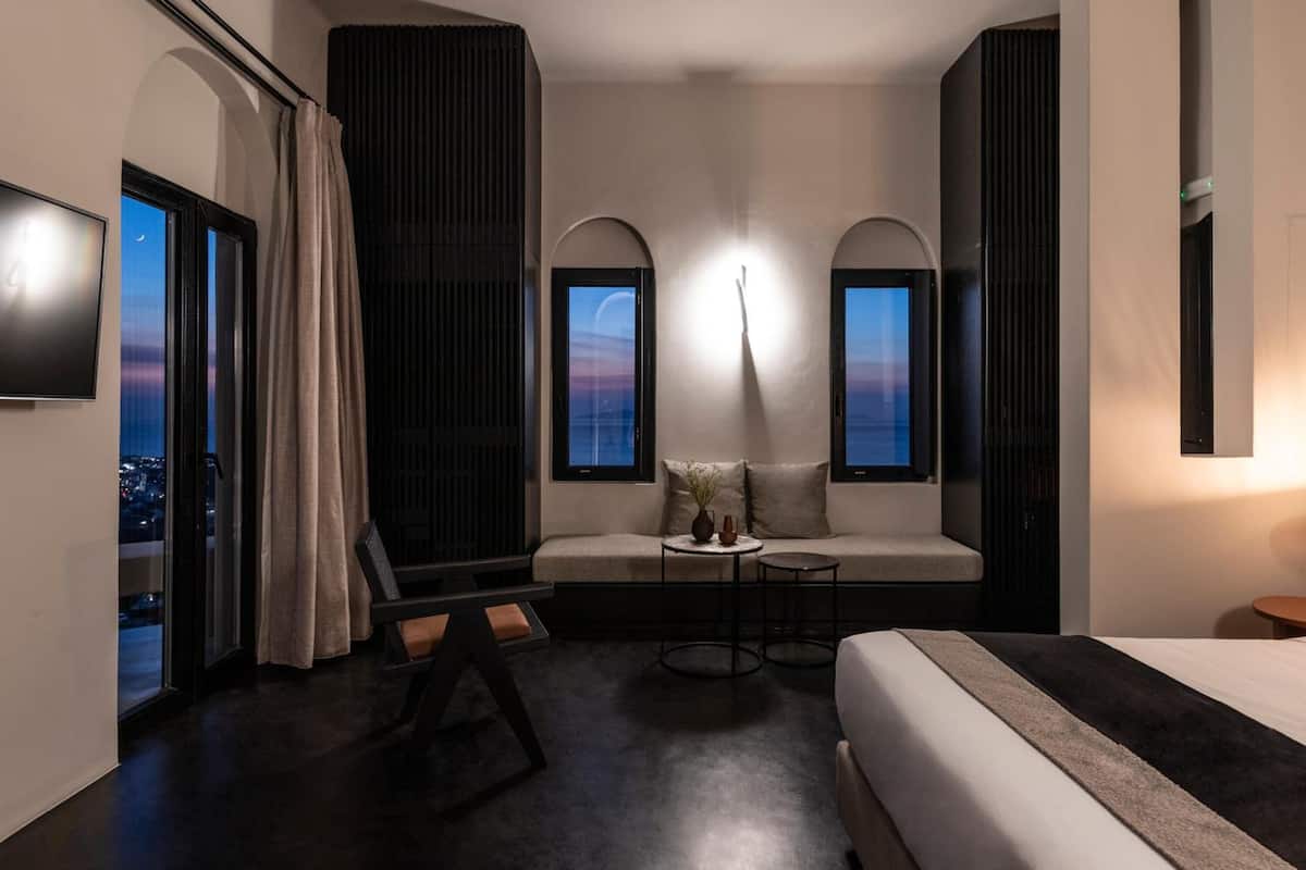 A modern hotel room with a large bed, a sofa, a small table, and two windows with views of the cityscape.