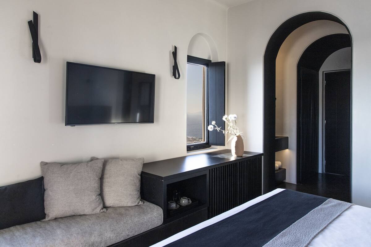 A modern bedroom with a large bed, a TV mounted on the wall, a console table with a vase of flowers, and a view through a window.