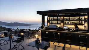 An outdoor bar area with a view of the ocean, tables, chairs, and a bar stocked with bottles. - Nimbus Santorini (Santorini)