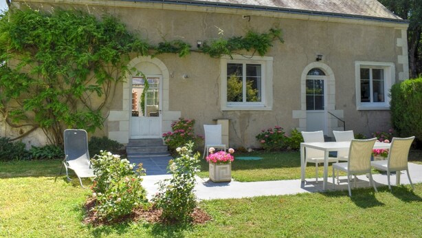 Une maison à deux étages avec un toit en pente, une porte d’entrée en arc de cercle et un petit porche agrémenté d’une table et de chaises.
