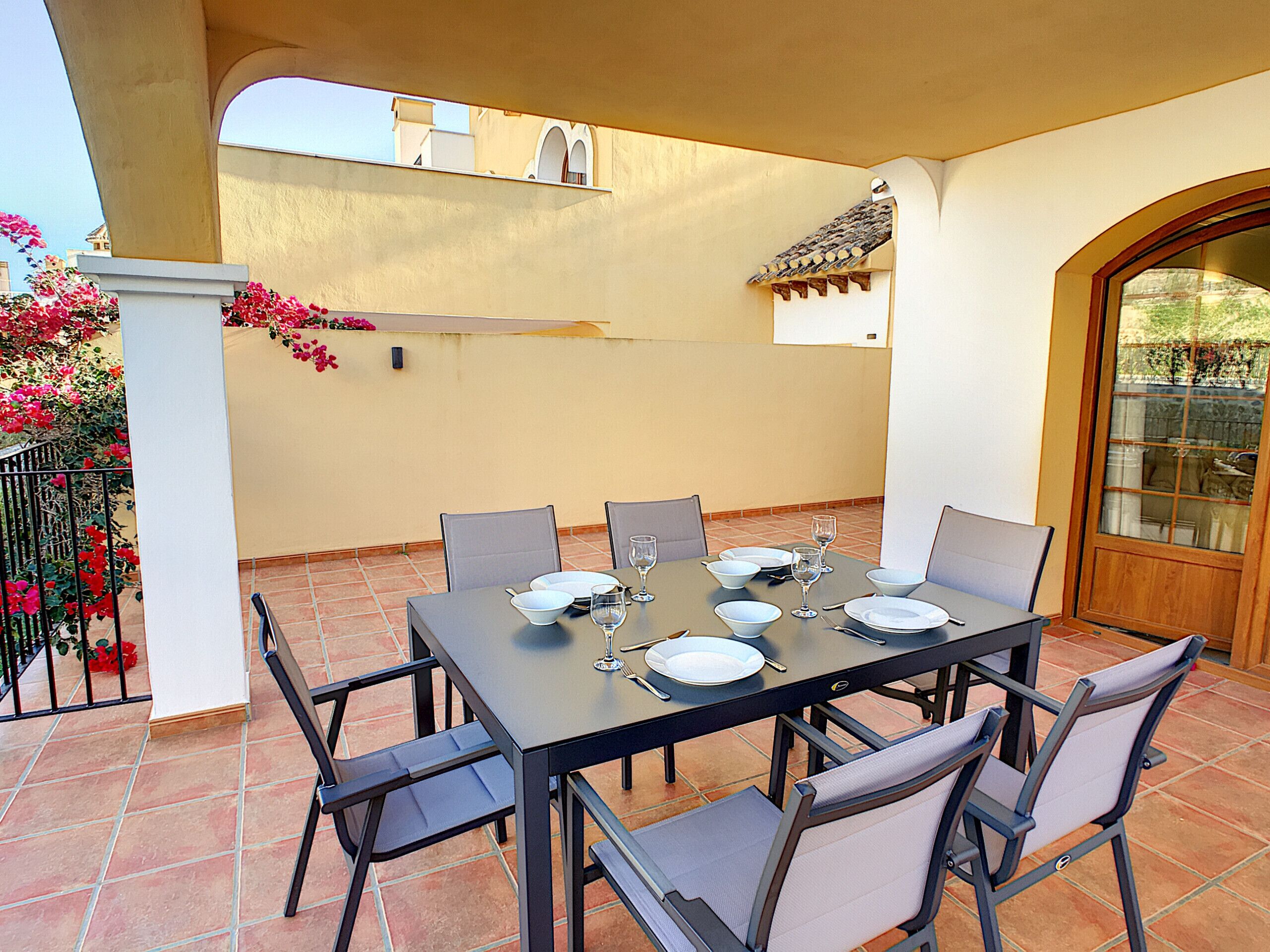 Un comedor con una mesa dispuesta para seis personas, rodeado de sillas, en un patio con baldosas y vistas a un edificio y un balcón con flores.