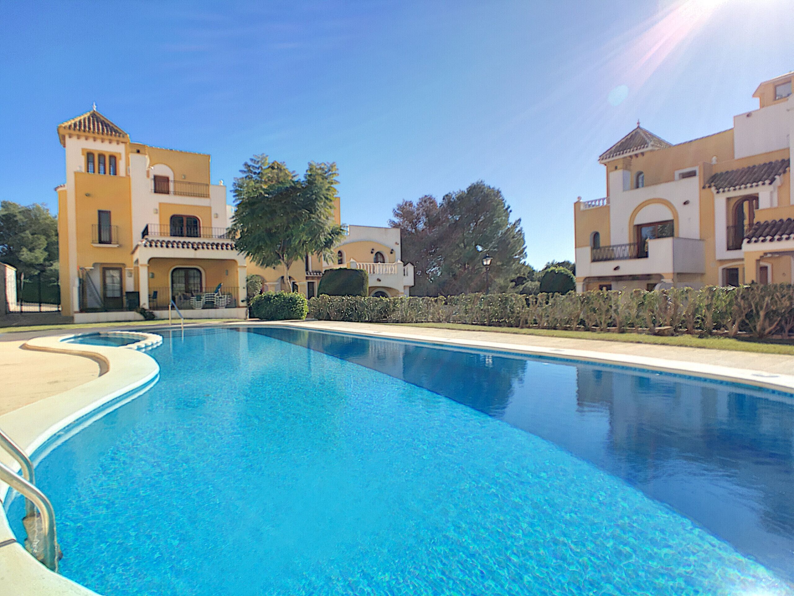Una piscina circondata da edifici dall'architettura mediterranea, cielo sereno e prato ben curato.