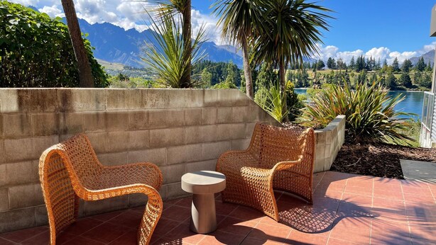 Een terras met rieten stoelen en een tafeltje, met uitzicht op een meer en bergen.