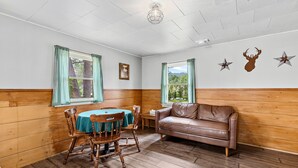 A room with wooden walls, a brown leather couch, a wooden dining table with chairs, and two windows with curtains.