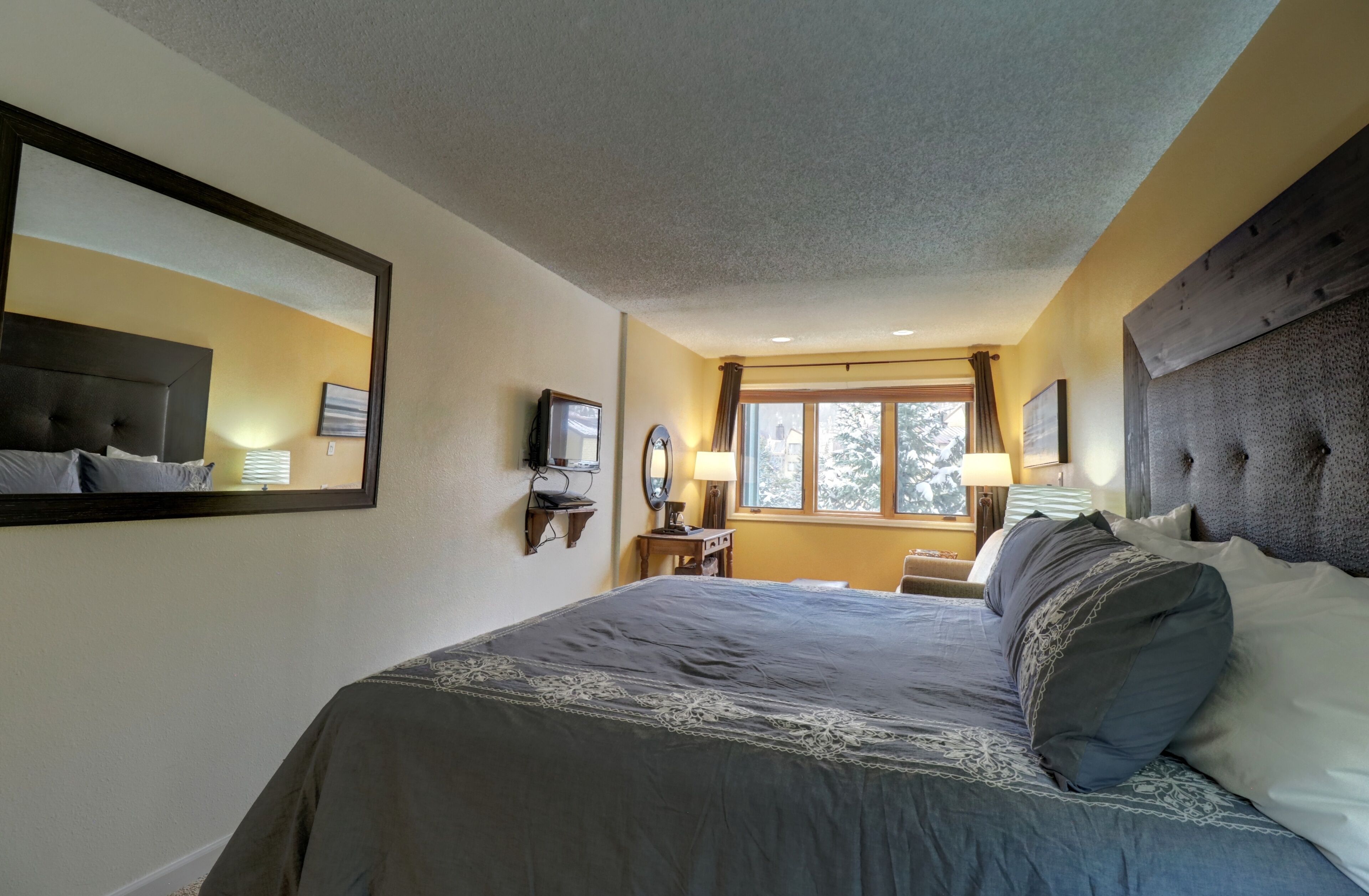 Une chambre à coucher avec un grand lit, une tête de lit, une table de chevet, un miroir et une fenêtre avec des rideaux.