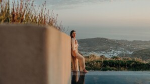 A person sitting by a pool with a view of hills and a cloudy sky. - Santorini Sky, Luxury Resort (Santorini)