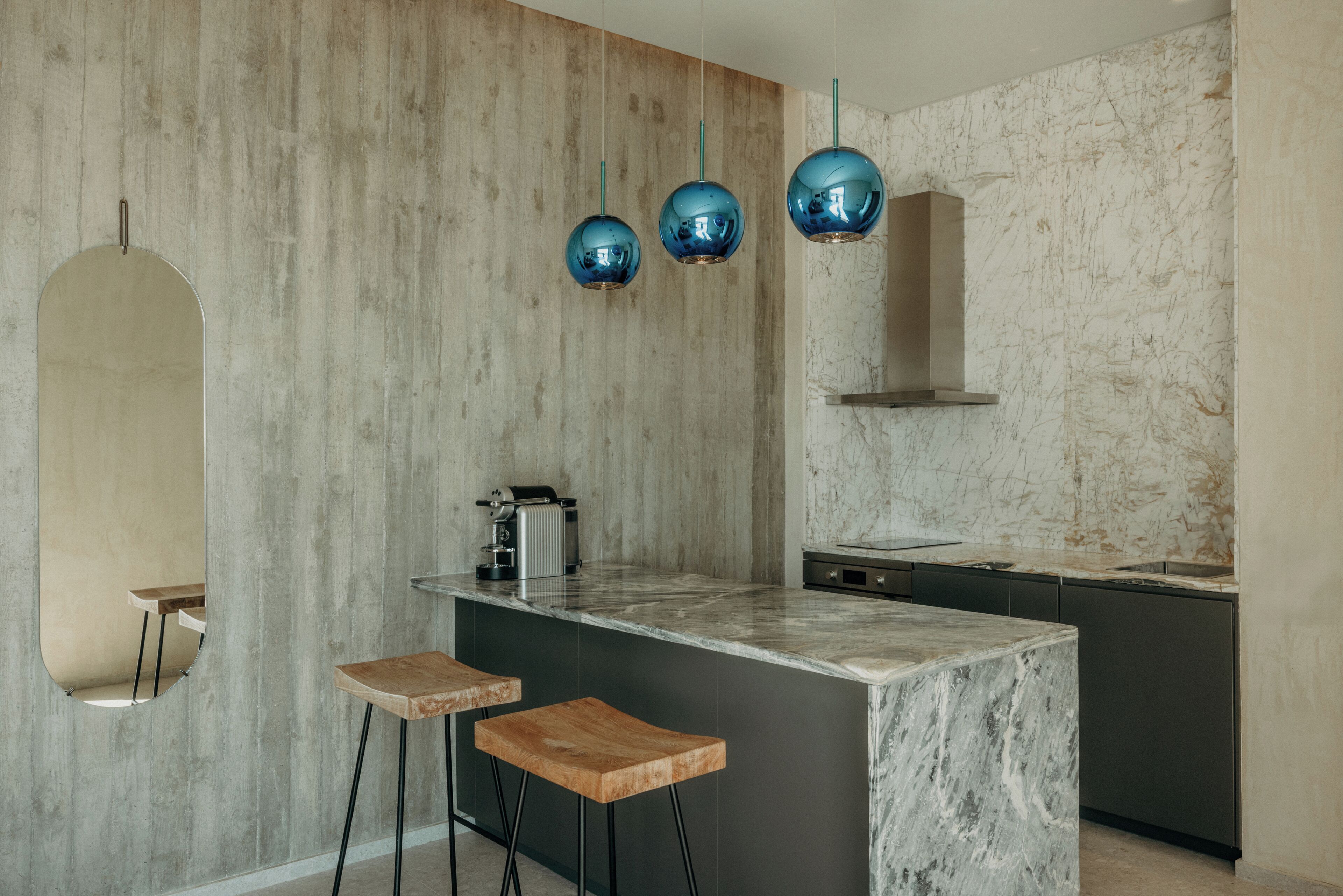 A modern kitchen with a marble countertop, a built-in oven, and a coffee machine. There are two wooden stools and three blue pendant lights.