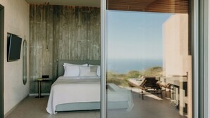 A modern bedroom with a large bed, a bedside table, and a TV. There is a balcony with outdoor furniture. - Santorini Sky, Luxury Resort (Santorini)
