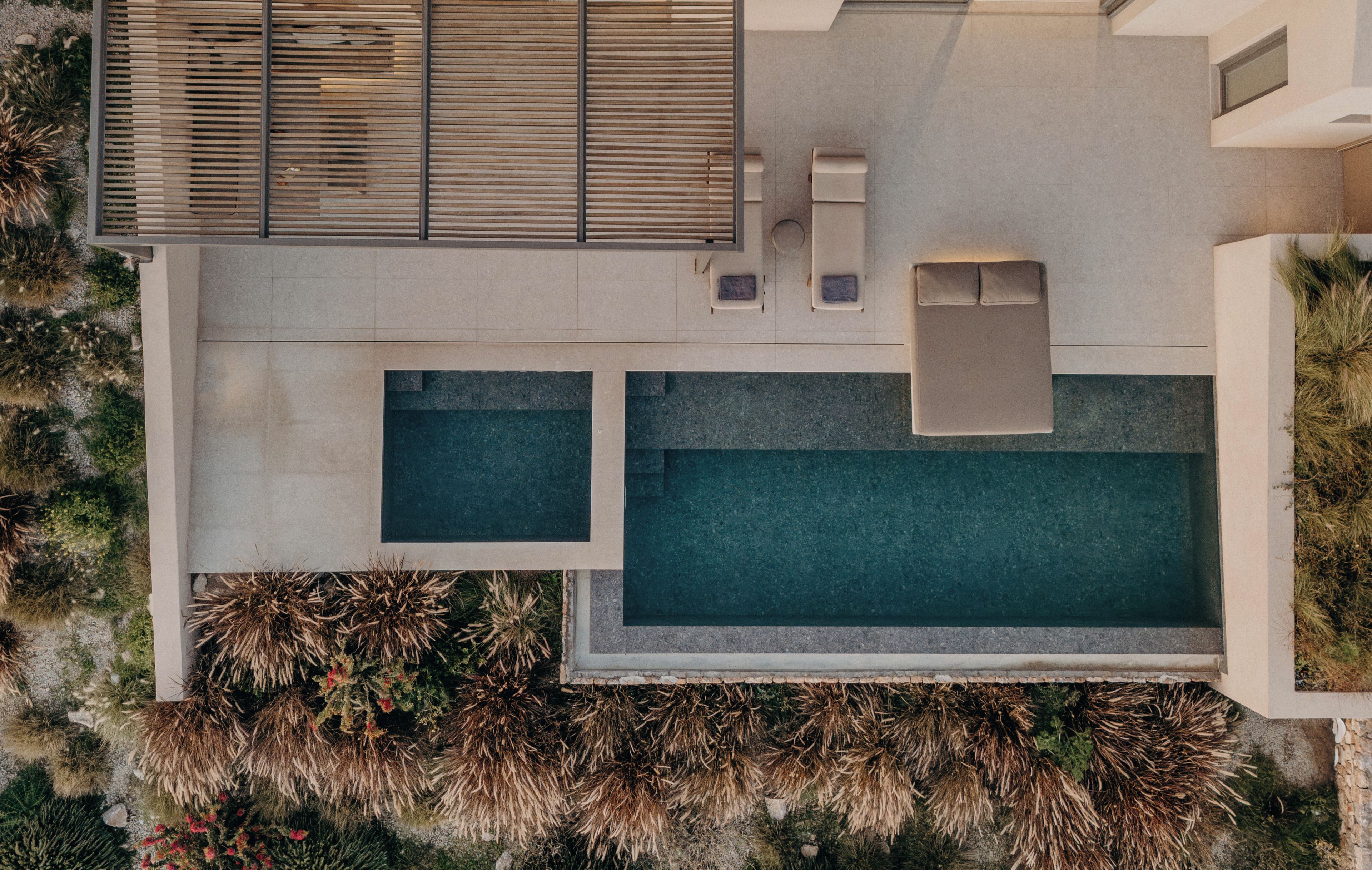 An aerial view of a modern outdoor pool area with two separate pools, a seating area with lounge chairs, and a wooden deck.