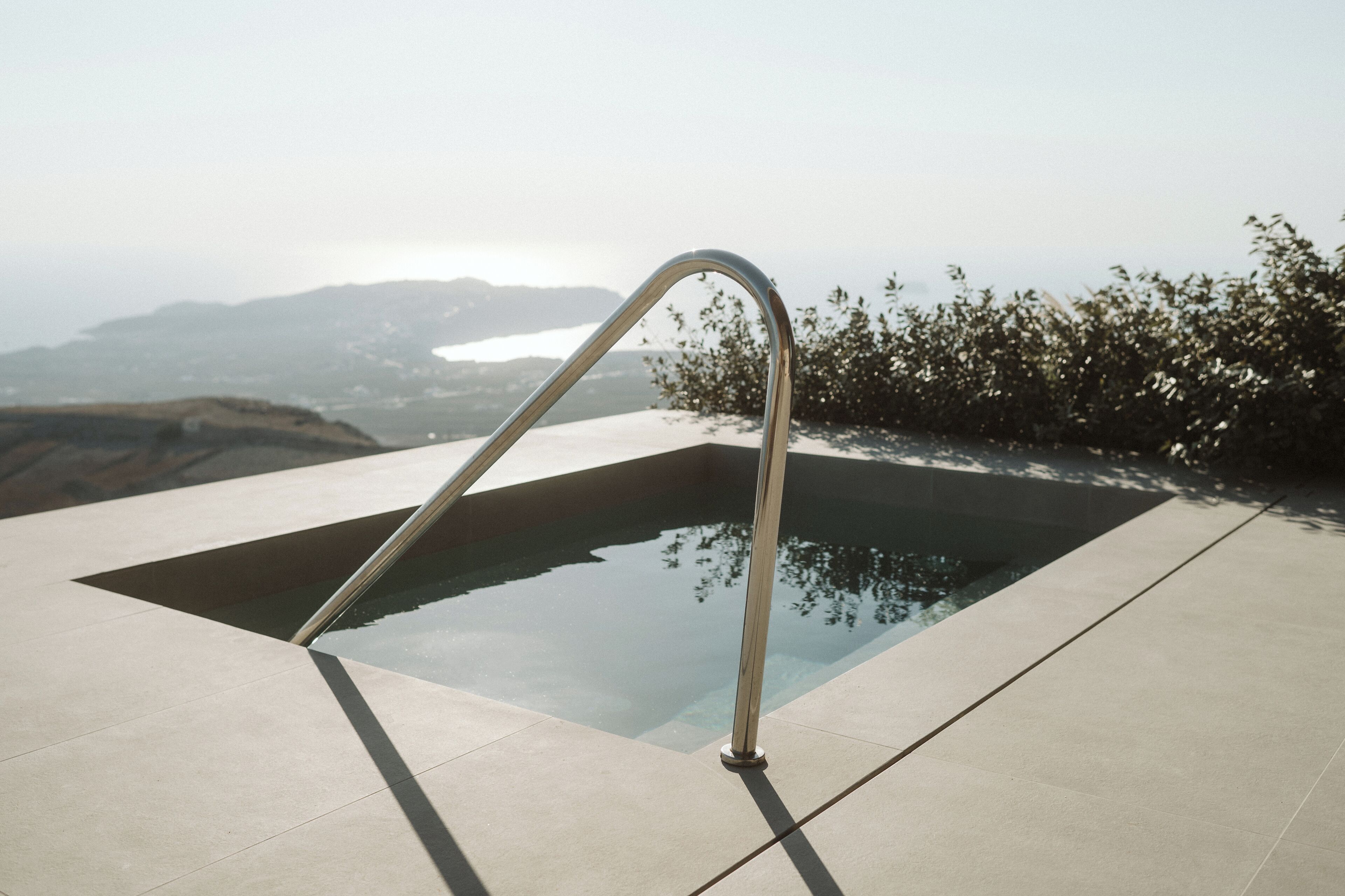 A modern outdoor pool with a metal ladder, surrounded by a concrete deck and overlooking a scenic landscape.