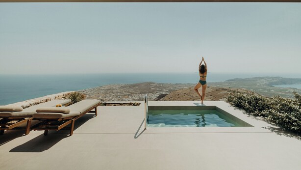 A pool with a view of the ocean, lounge chairs, and a person doing yoga. - Santorini Sky, Luxury Resort (Santorini)